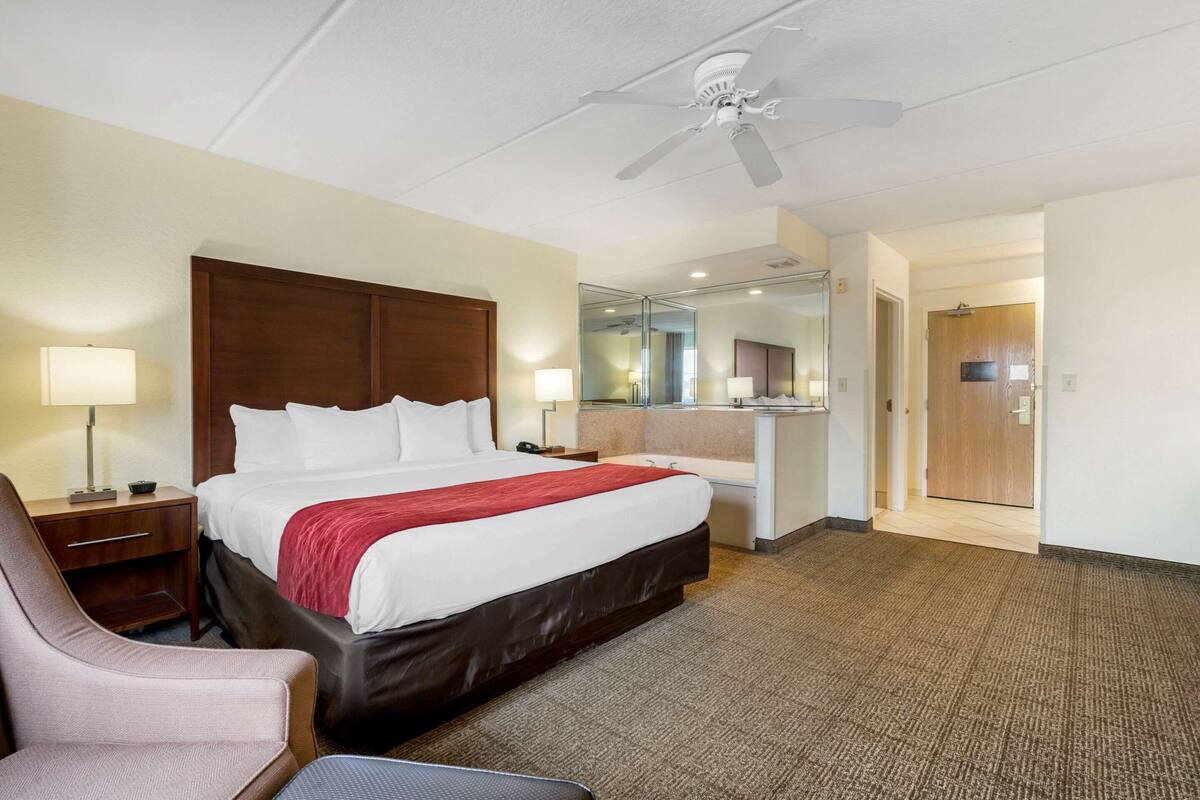 A hotel room with a large bed, a desk with a lamp, a chair, a ceiling fan, and a bathroom visible through a glass door.
