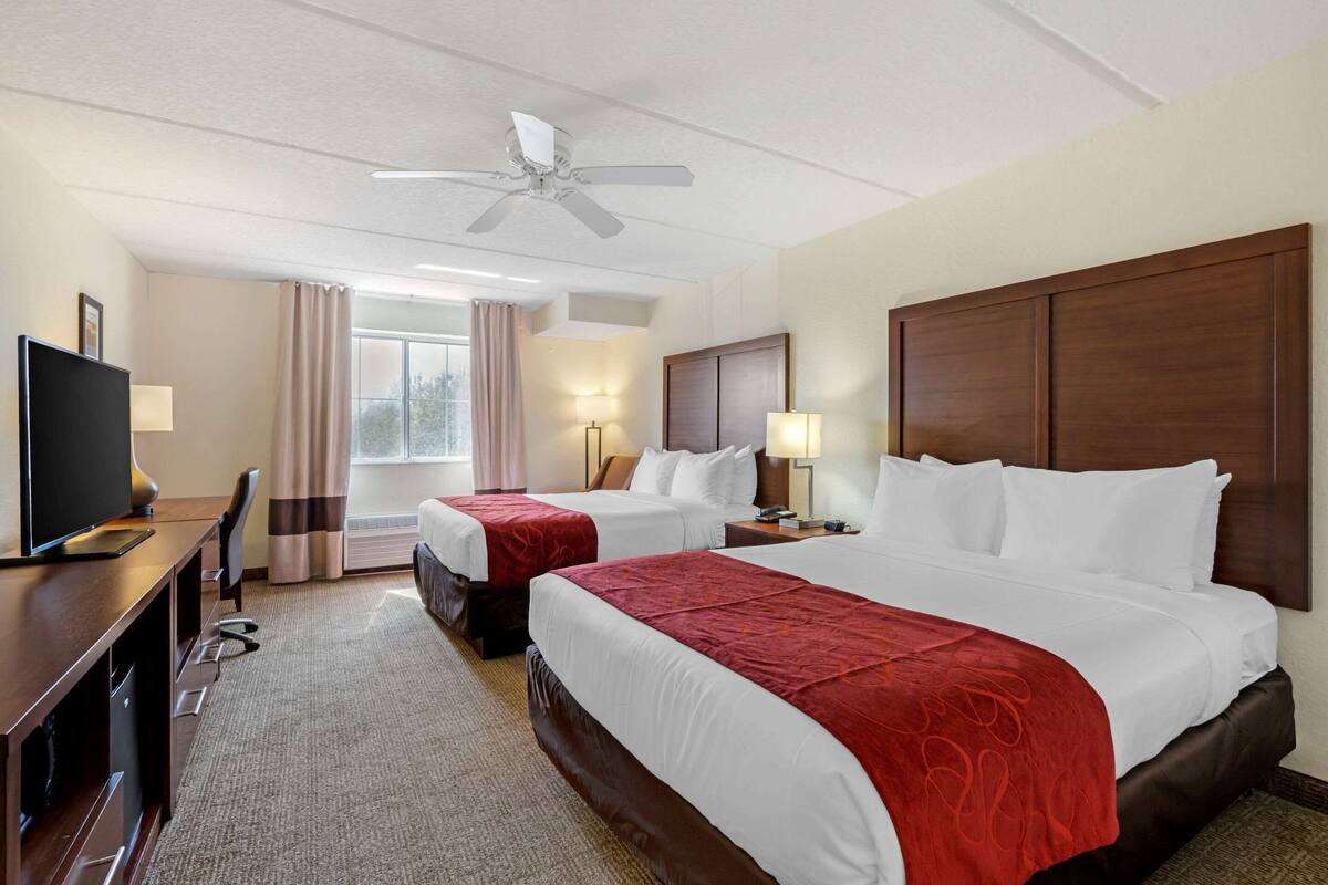 A hotel room with two beds, a desk, a TV, and a ceiling fan.