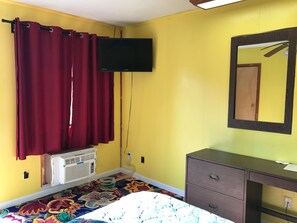 A room with yellow walls, a bed with a colorful floral bedspread, a dark wooden dresser, a mirror, a flat-screen TV mounted on the wall, and a window with red curtains.