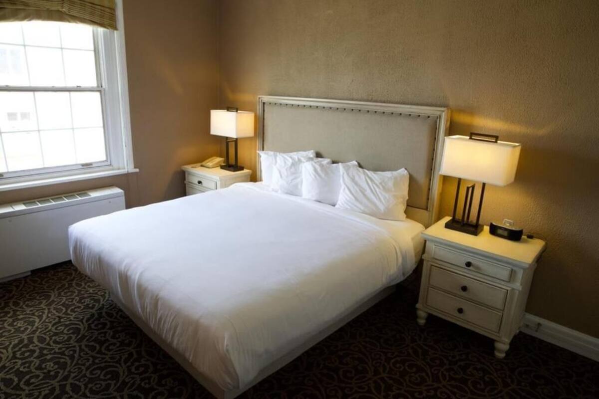 A neatly made bed with white linens, two bedside tables with lamps, a window with blinds, and a patterned carpet.