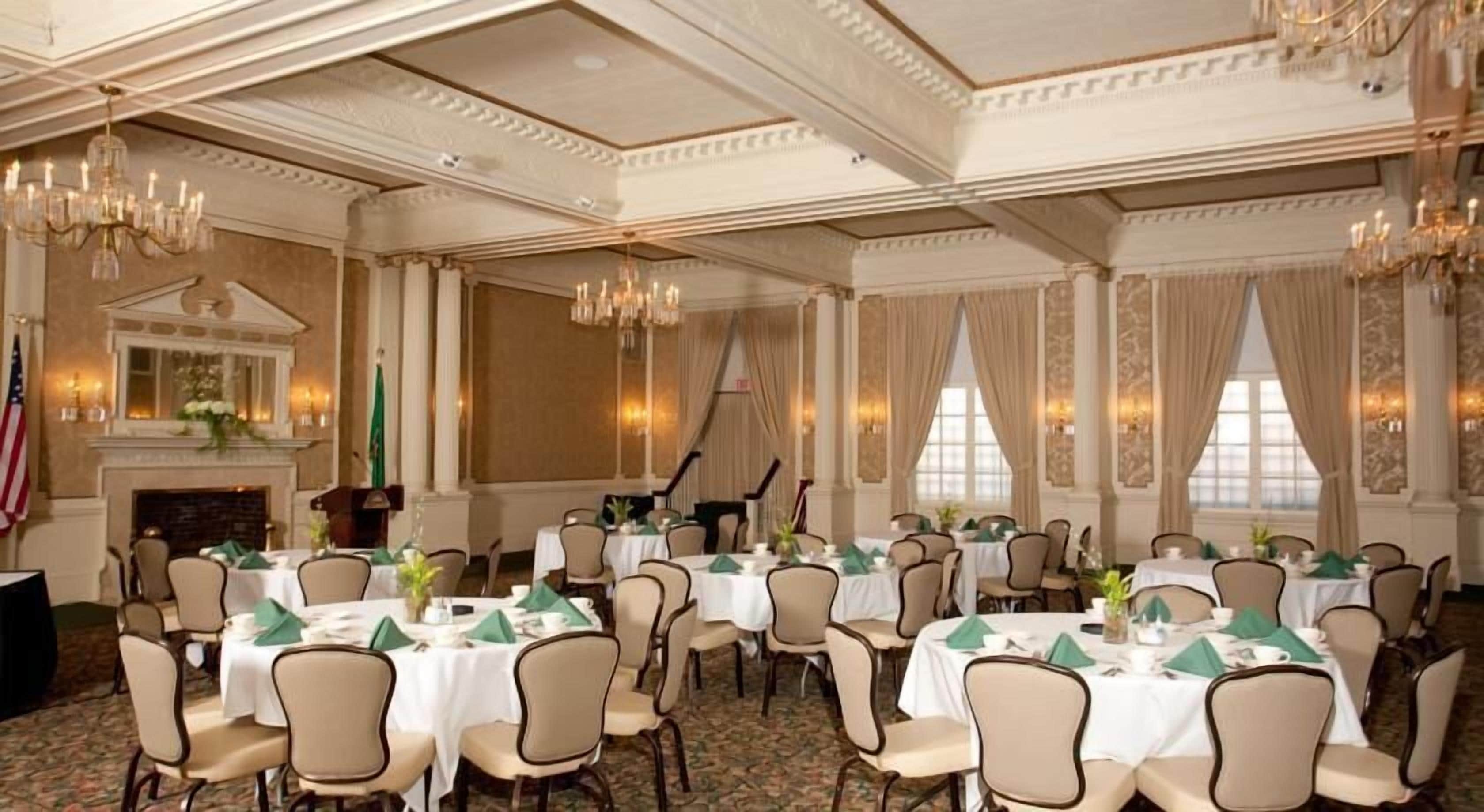 A formal dining room with multiple round tables, chairs, chandeliers, and a fireplace.