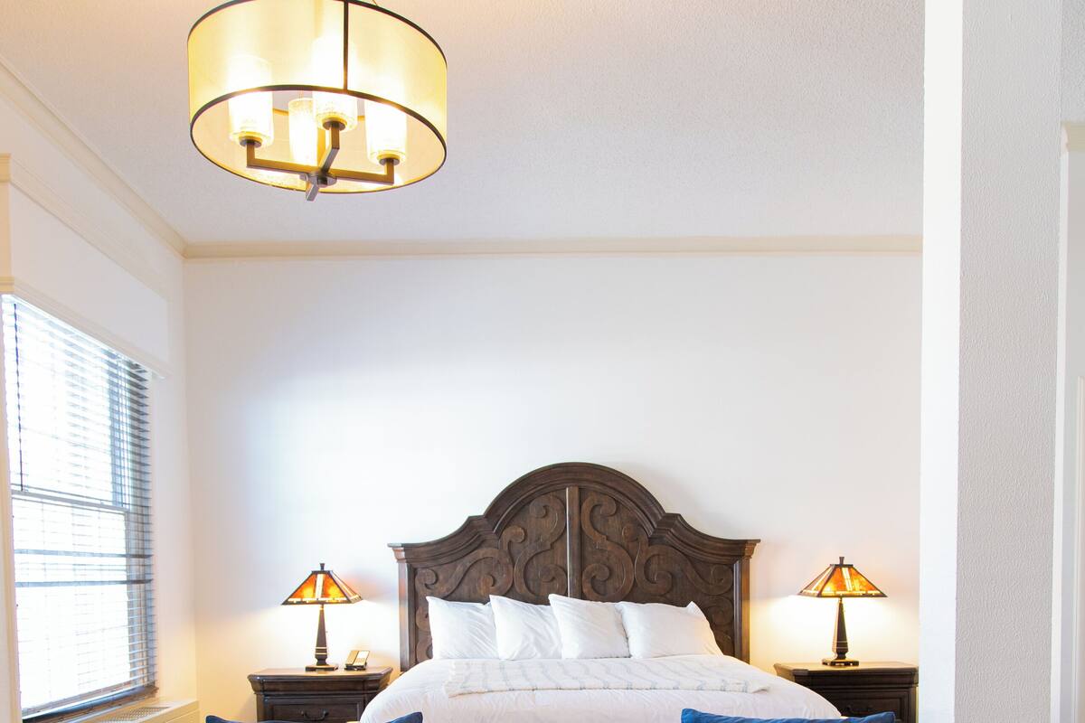 A bedroom with a large bed, two bedside lamps, a blue sofa, a wooden coffee table, and a vase with flowers.