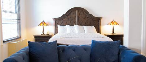 Un dormitorio con una cama grande, dos lámparas de noche, un sofá azul, una mesa de centro de madera y un florero con flores.