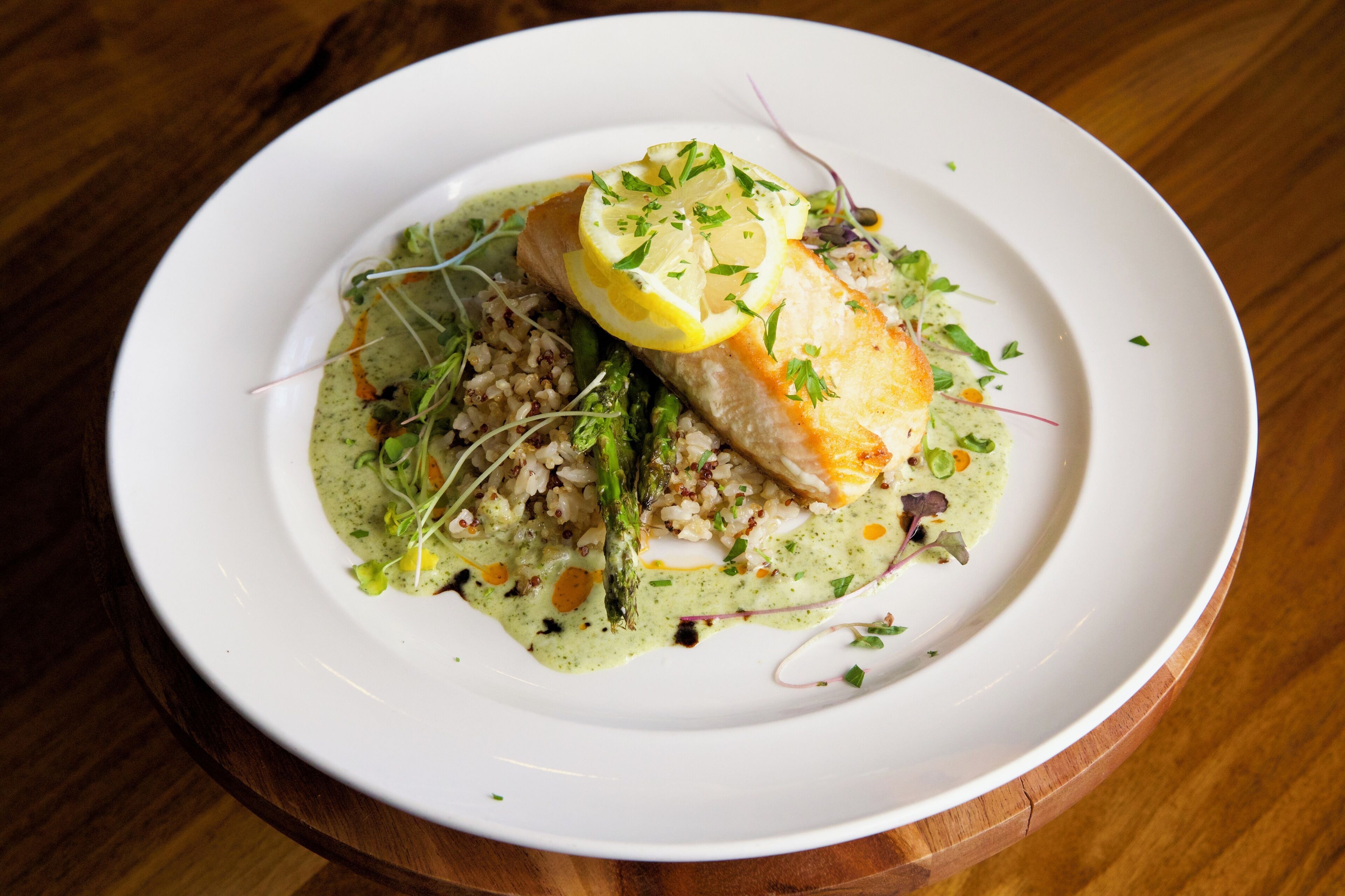 Un plato blanco con un filete de pescado, espárragos y una salsa verde sobre una mesa de madera.