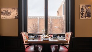 Mesa de jantar posta para dois, com vista para um edifício de tijolos através de uma grande janela.