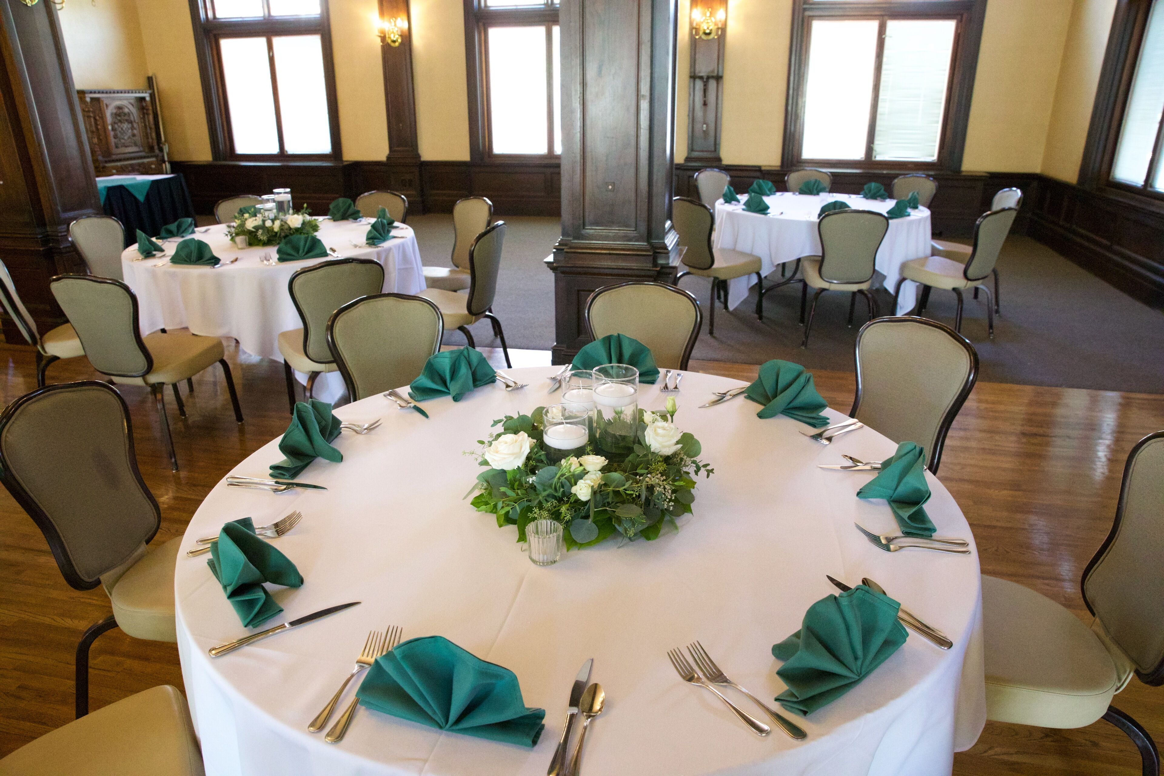A banquet hall with round tables set for a formal event, featuring green napkins, white tablecloths, and floral centerpieces.