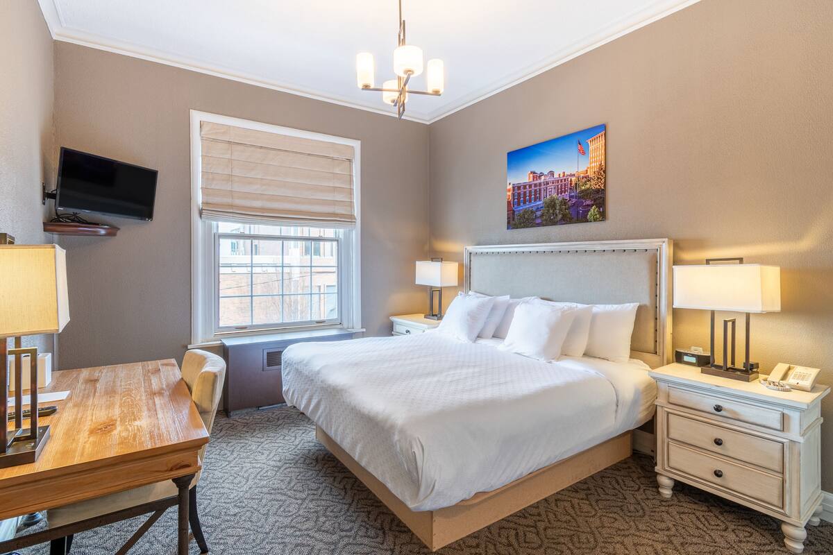 A hotel room with a bed, bedside tables, a desk, a chair, a TV mounted on the wall, and a window with blinds.