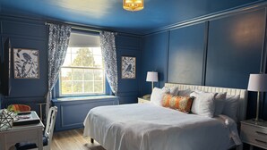 Quarto com uma cama grande, uma janela com cortinas estampadas, uma escrivaninha com cadeira e uma mesa de cabeceira com abajur.