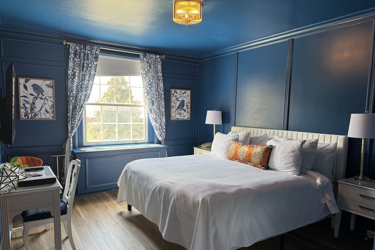 A bedroom with a large bed, a window with patterned curtains, a desk with a chair, and a nightstand with a lamp.