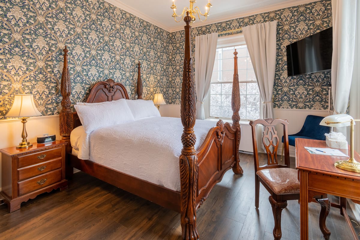 A four-poster bed with a wooden frame, a nightstand with drawers, a desk with a lamp, and a chair.