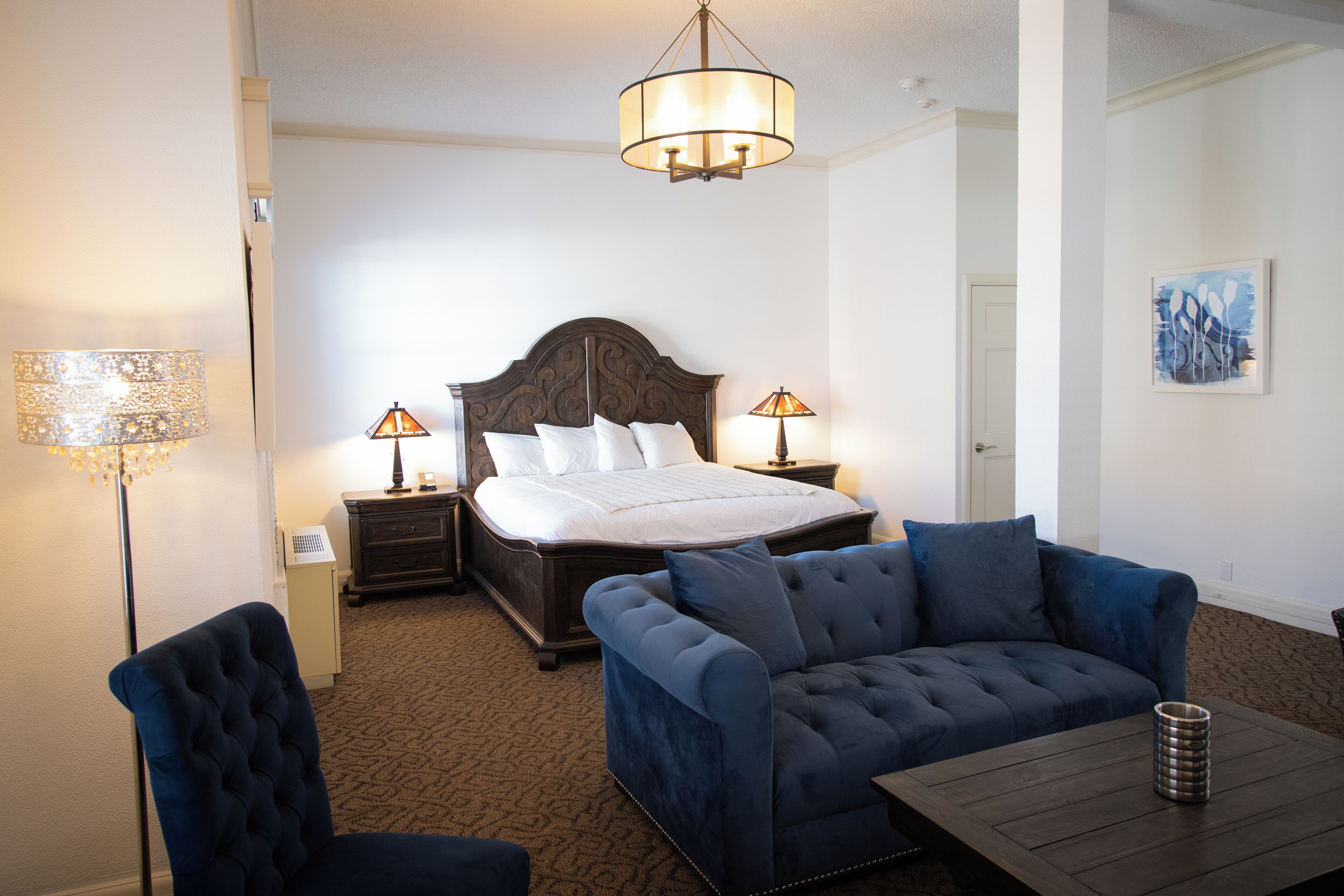 Quarto de hotel com uma cama grande, duas luminárias de cabeceira, um sofá azul escuro e uma mesa de centro de madeira.