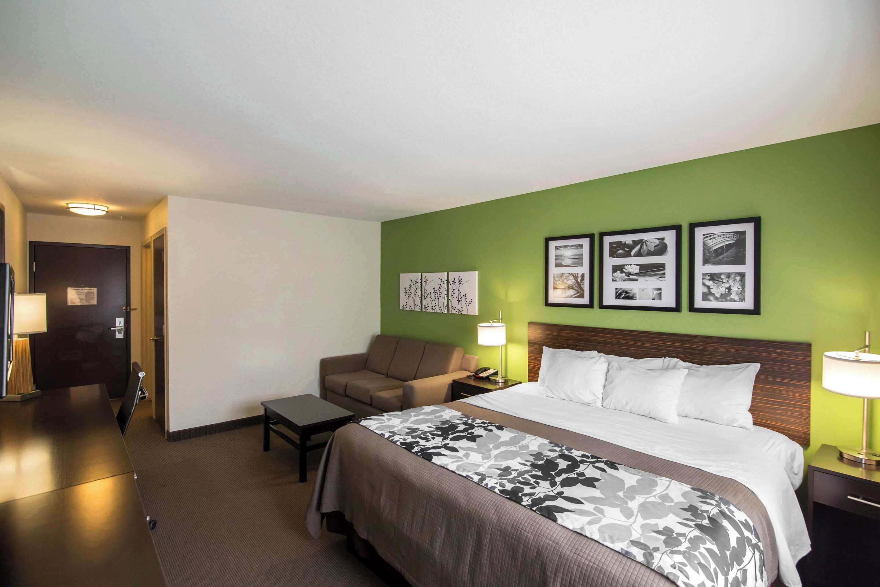 Habitación de hotel con cama, sofá, escritorio y mesita de noche. En la pared hay cuadros enmarcados y una puerta al fondo.