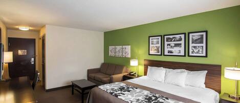 A hotel room with a bed, a sofa, a desk, and a nightstand. There are framed pictures on the wall and a door in the background.