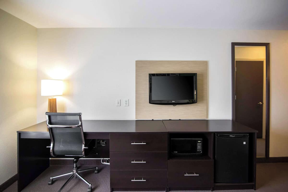 A hotel room with a dark wooden desk, a black office chair, a flat-screen TV mounted on the wall, a microwave, and a refrigerator.
