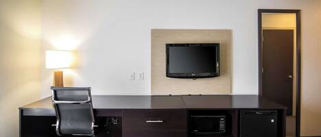 A hotel room with a dark wooden desk, a black office chair, a flat-screen TV mounted on the wall, a microwave, and a refrigerator.