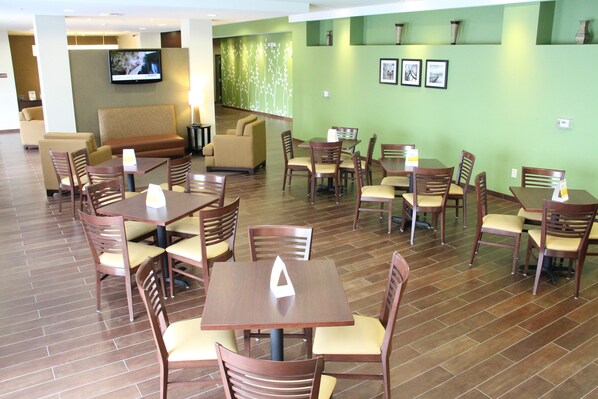 A hotel lobby with multiple seating areas including sofas and tables, a flat-screen TV mounted on the wall, and a green wall with framed pictures.
