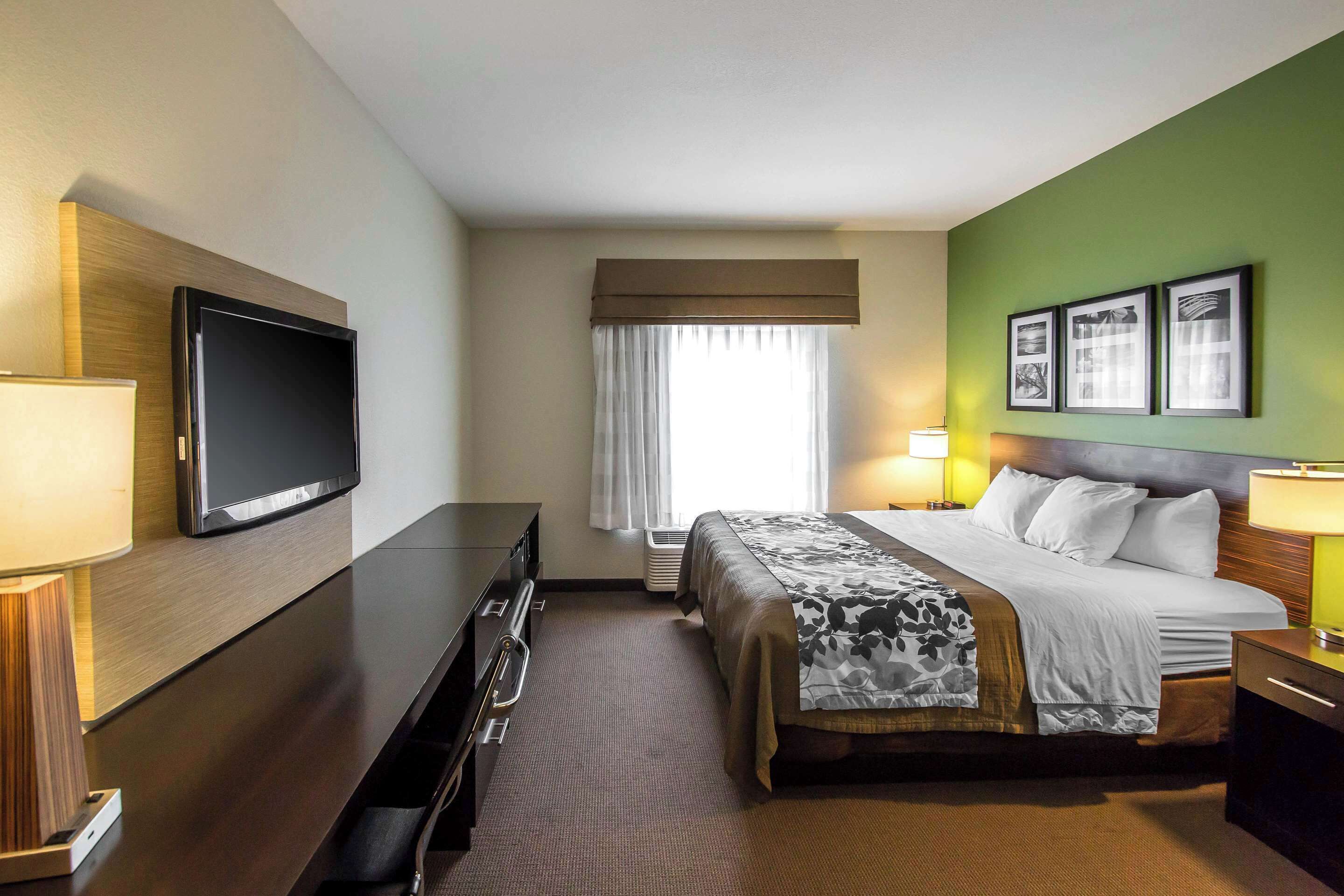 Quarto de hotel com cama, televisão, escrivaninha e janela com cortinas.