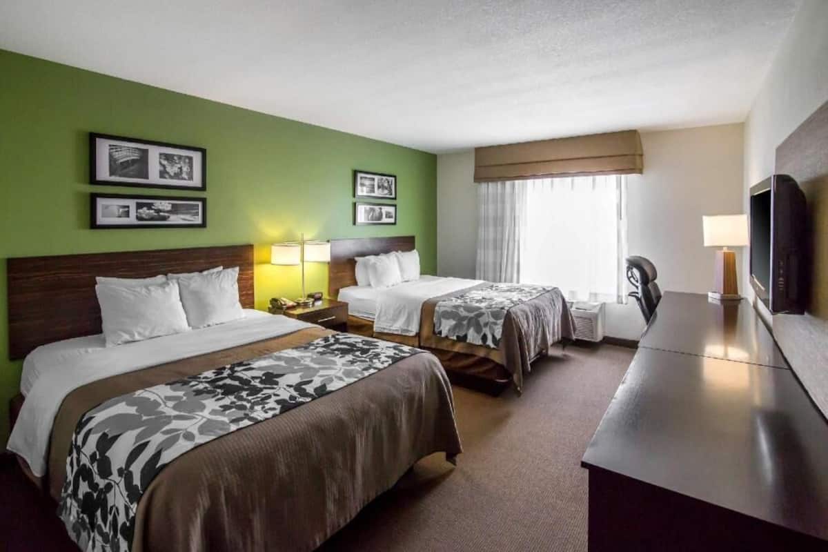 A hotel room with two beds, a desk, a TV, and framed pictures on the wall.
