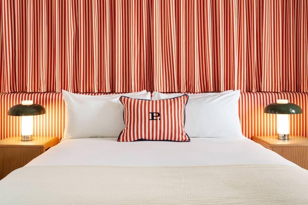 A hotel room with a bed, striped curtains, two bedside lamps, and a pillow with a letter 'P'.