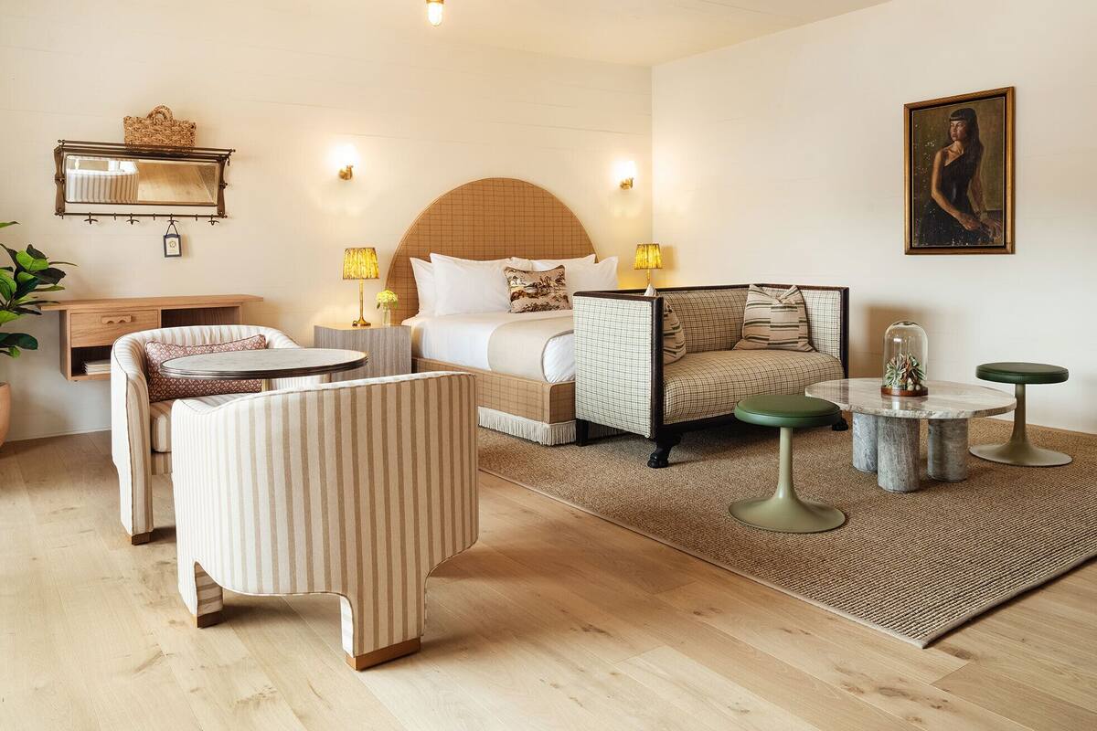 A hotel room with a bed, a sofa, a small table, and a chair. There is a lamp on the bedside table and a plant on the floor.