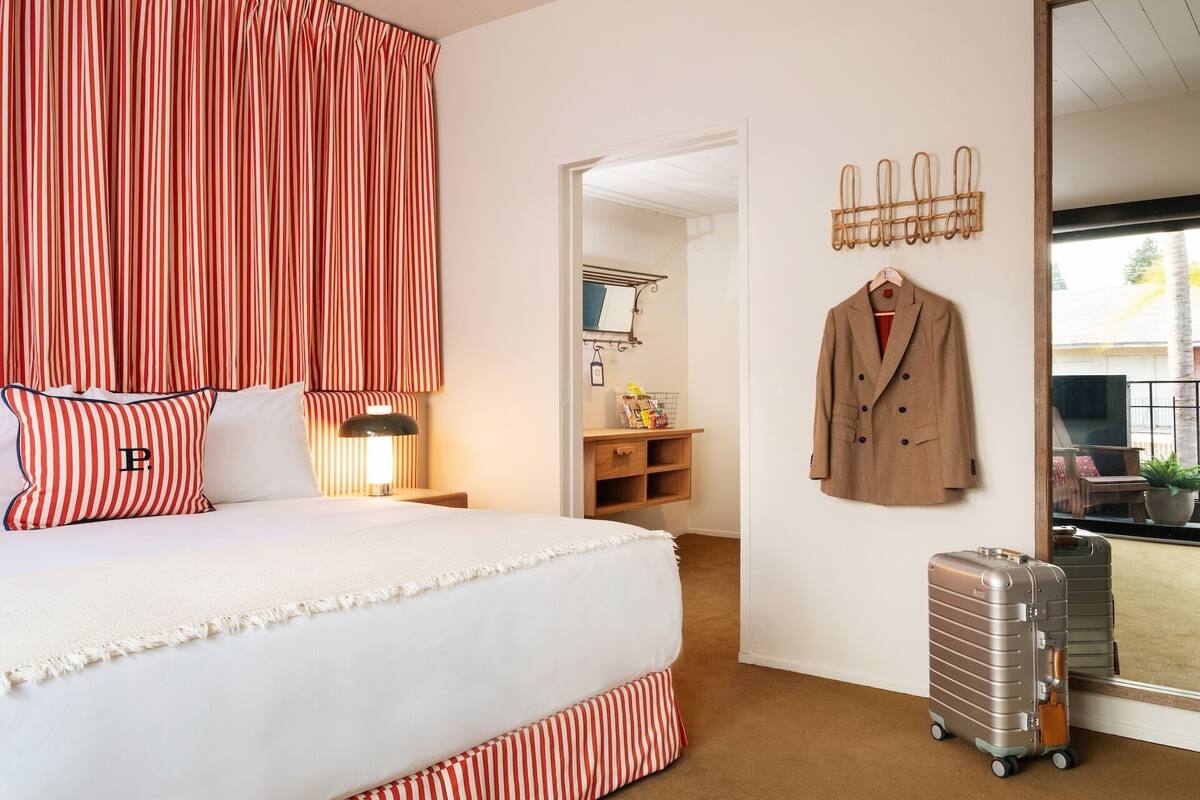 A hotel room with a bed, striped curtains, a coat rack with a coat, and a suitcase.
