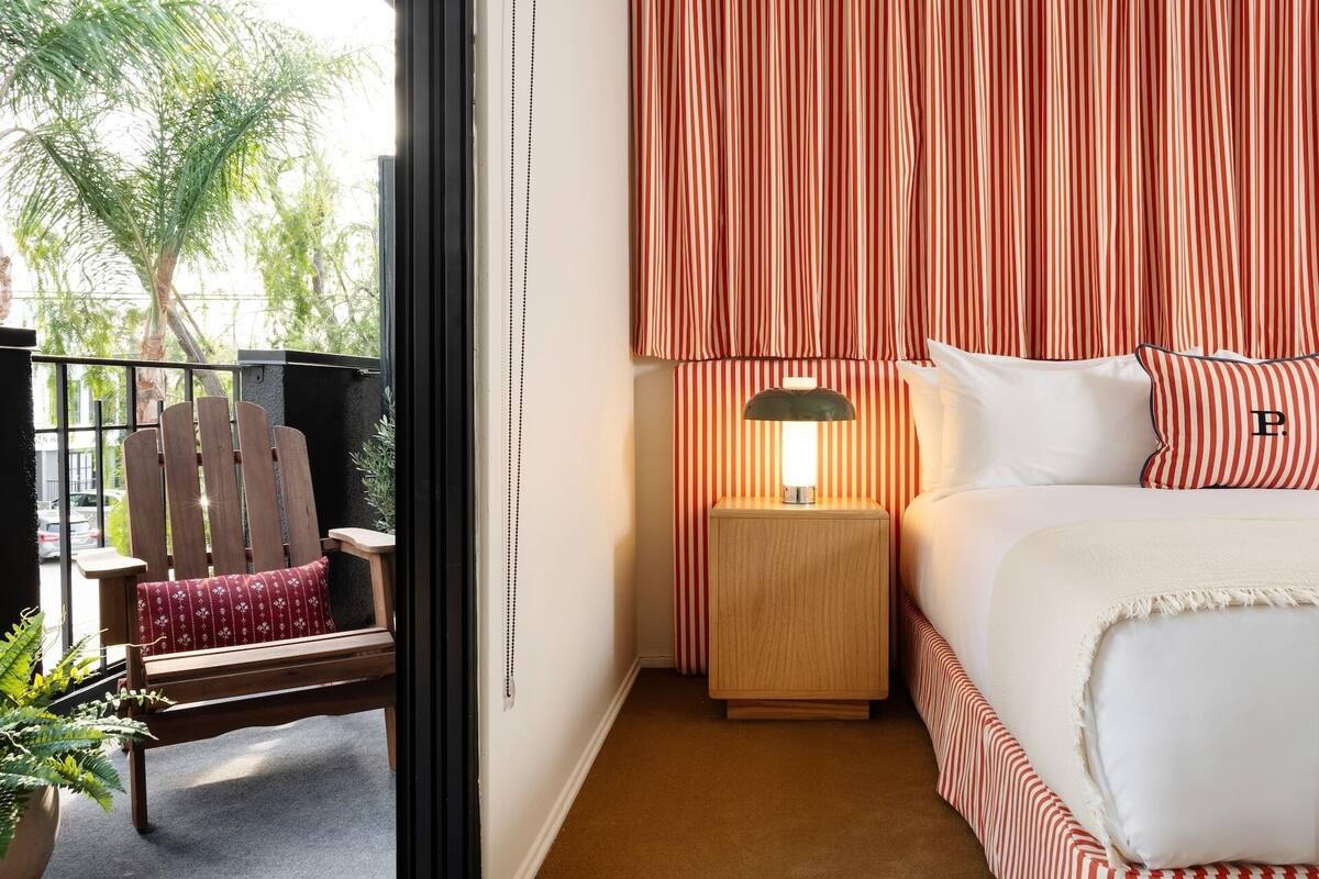 A hotel room with a bed, a nightstand with a lamp, a chair, and a view of the outdoors.