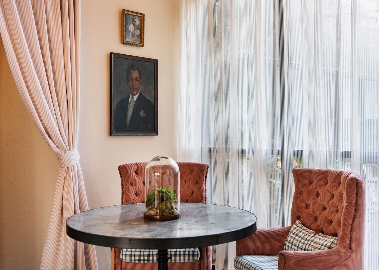 Una habitación con una mesa redonda, una cúpula de cristal con plantas, un retrato enmarcado y una ventana con cortinas translúcidas.