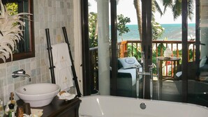 A bathroom with a large bathtub, a sink, and a mirror. There is a glass door leading to a balcony with a view of the ocean. - Dhevatara Beach Hotel & Spa (Praslin Island)