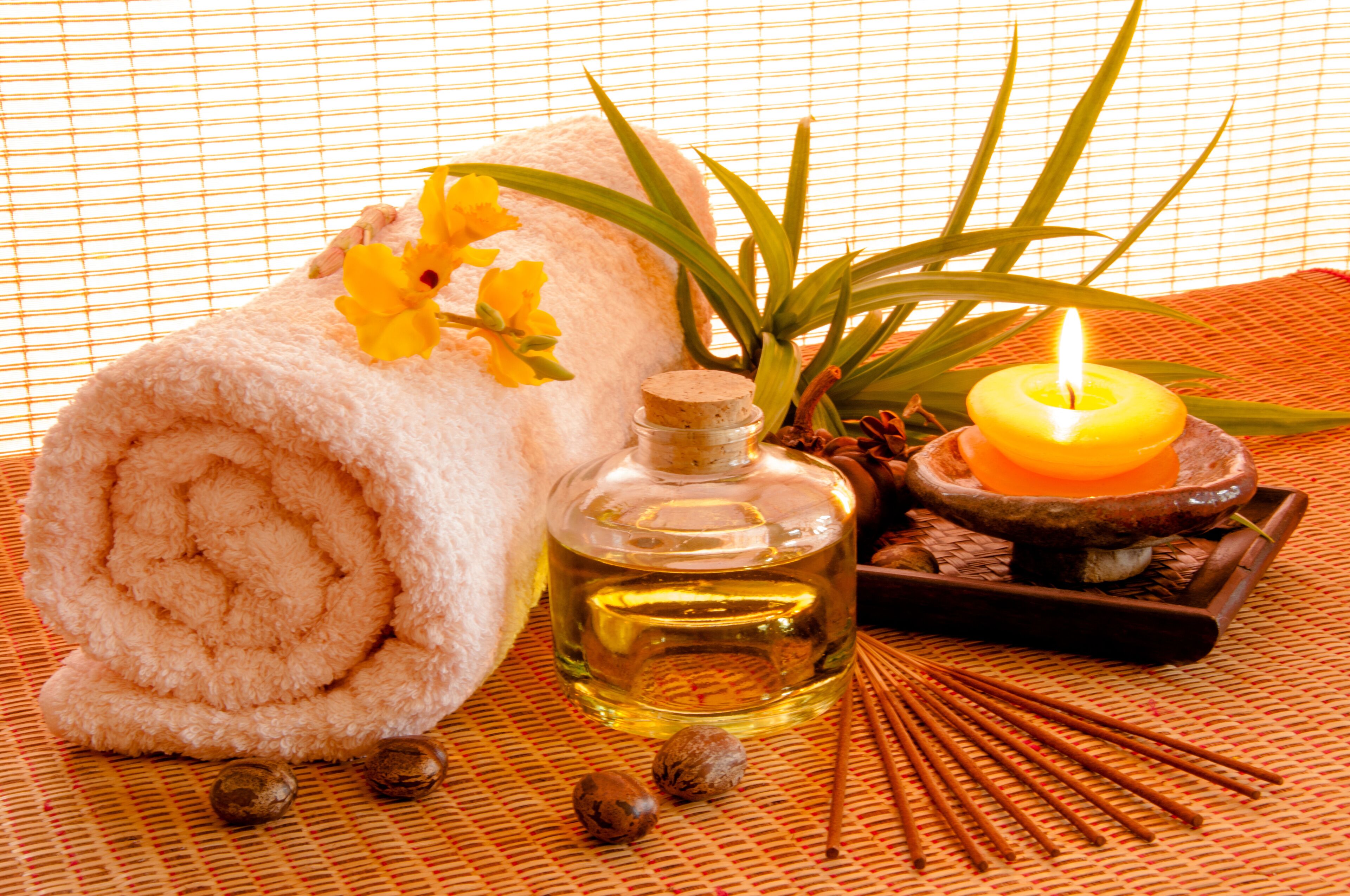 A rolled towel, a bottle of oil, a lit candle, and some incense sticks on a textured mat.