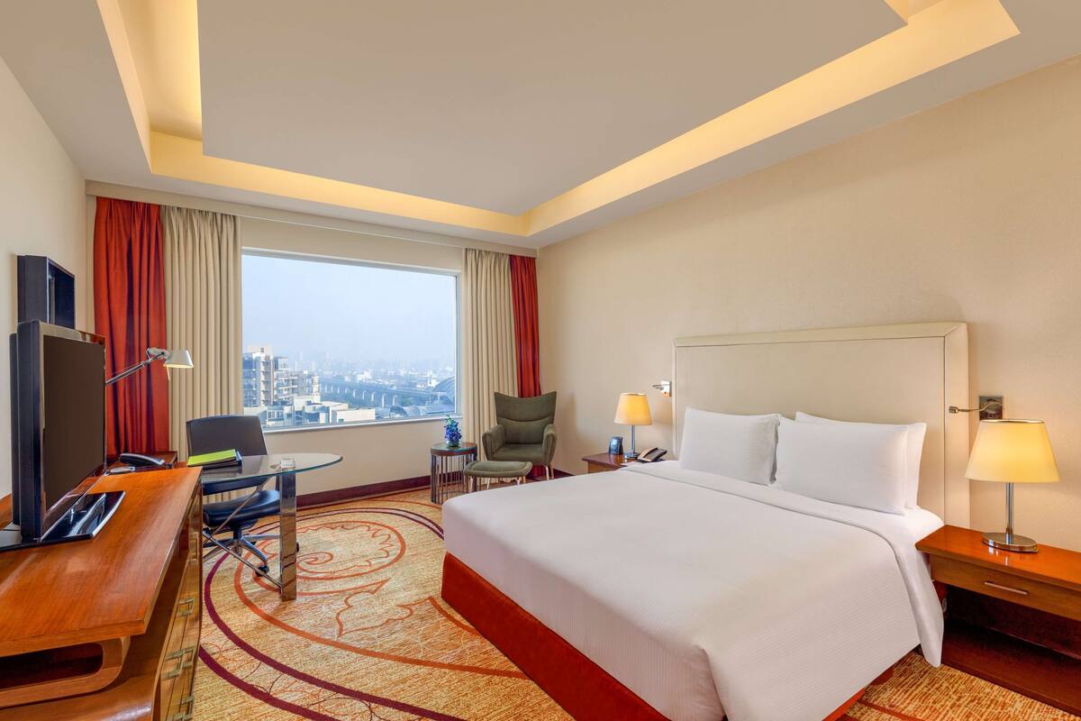 A hotel room with a large bed, a desk with a computer, a chair, a TV, and a view of the city.