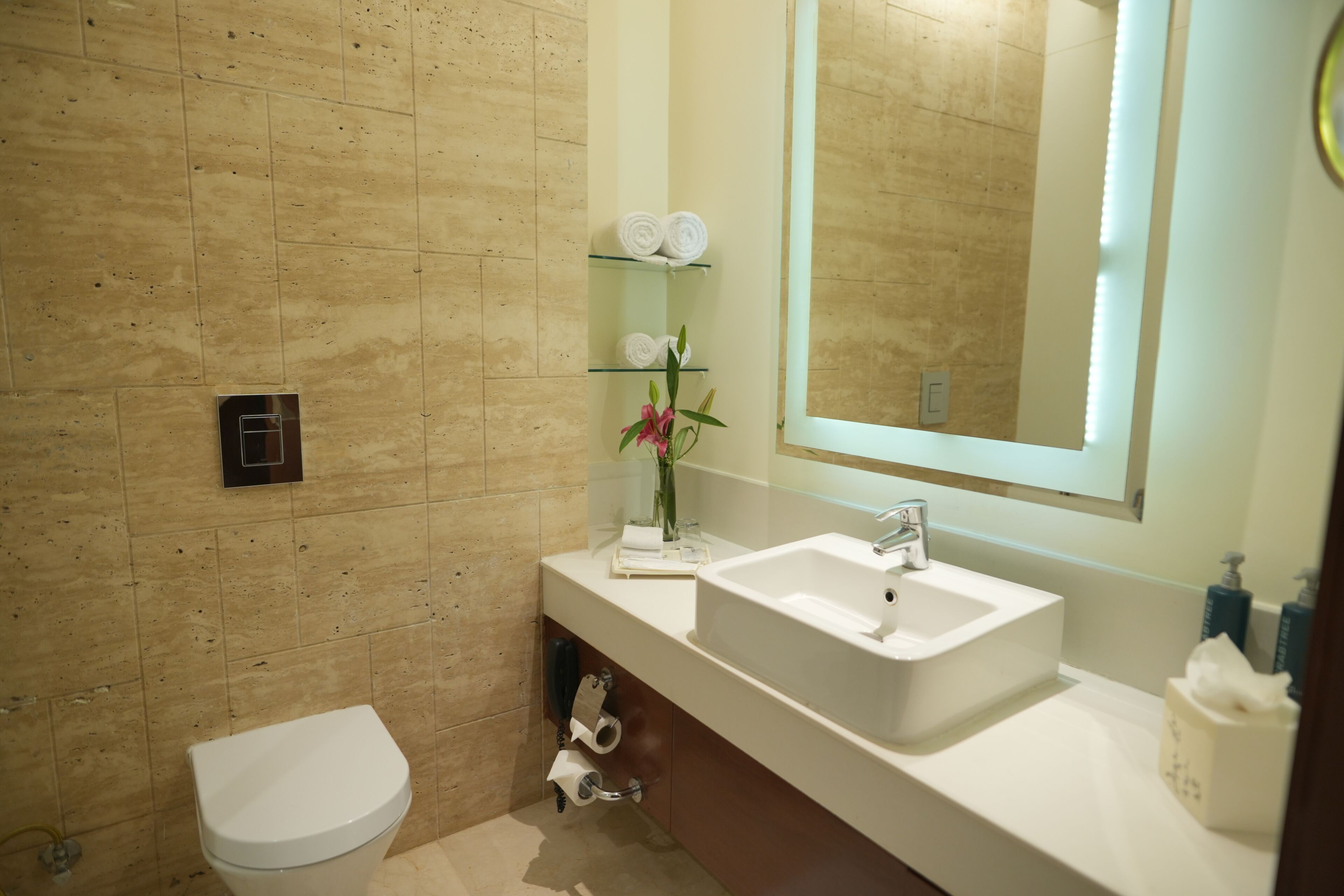 Une salle de bain avec un lavabo blanc, un miroir et des toilettes. On y trouve des serviettes sur une étagère et un vase avec des fleurs.