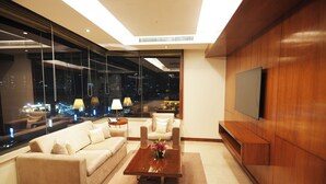 A modern living room with a sofa, coffee table, and TV against a wall with a large window overlooking a cityscape at night.