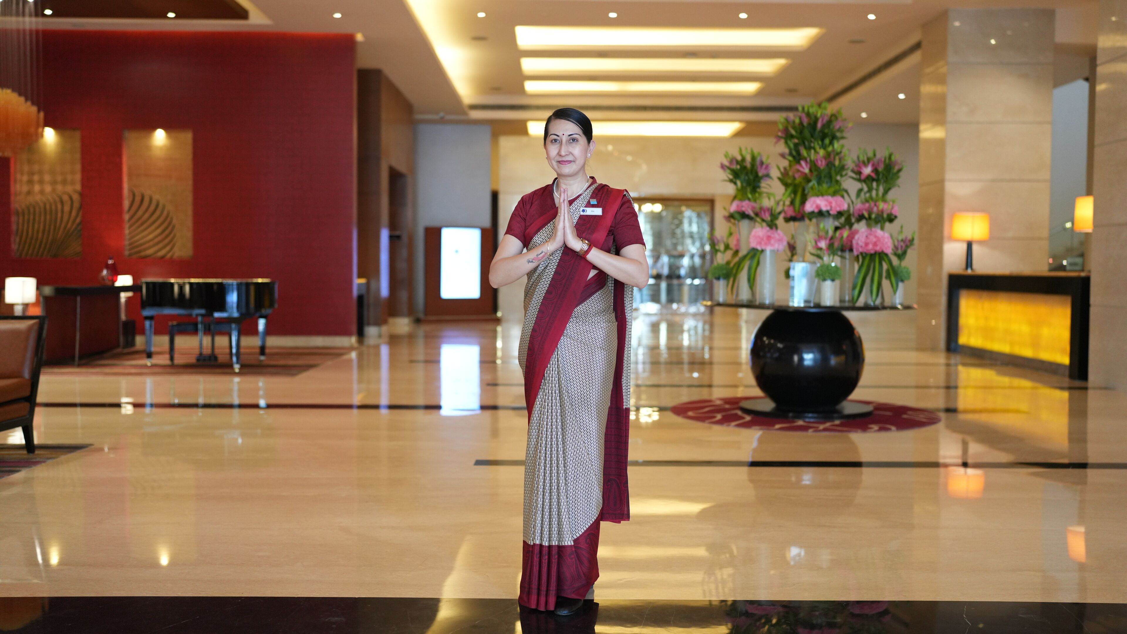 A woman in traditional attire standing in a modern lobby with a grand piano, a reception desk, and decorative floral arrangements.