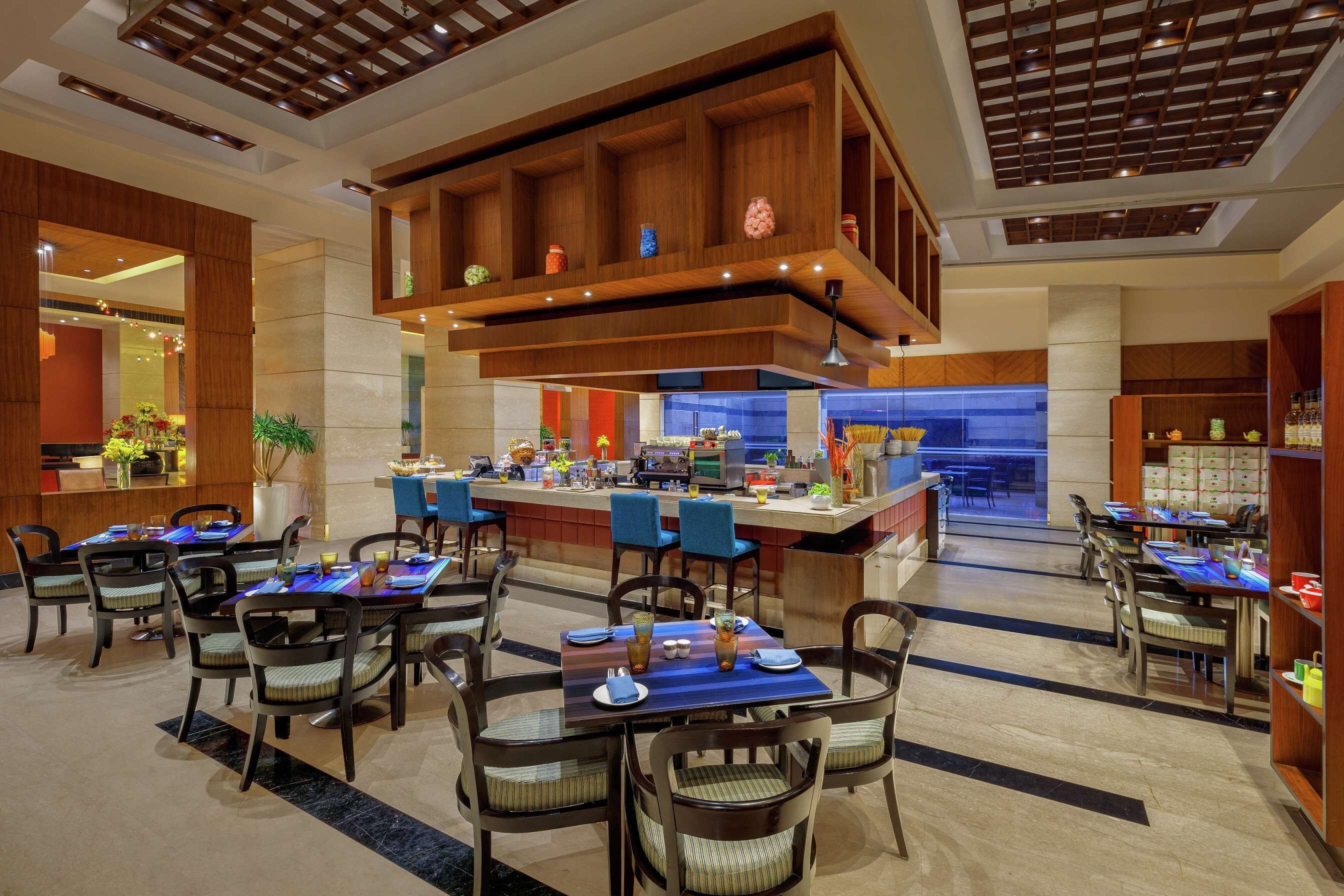 A modern hotel lobby with multiple dining tables, chairs, and a central bar area.