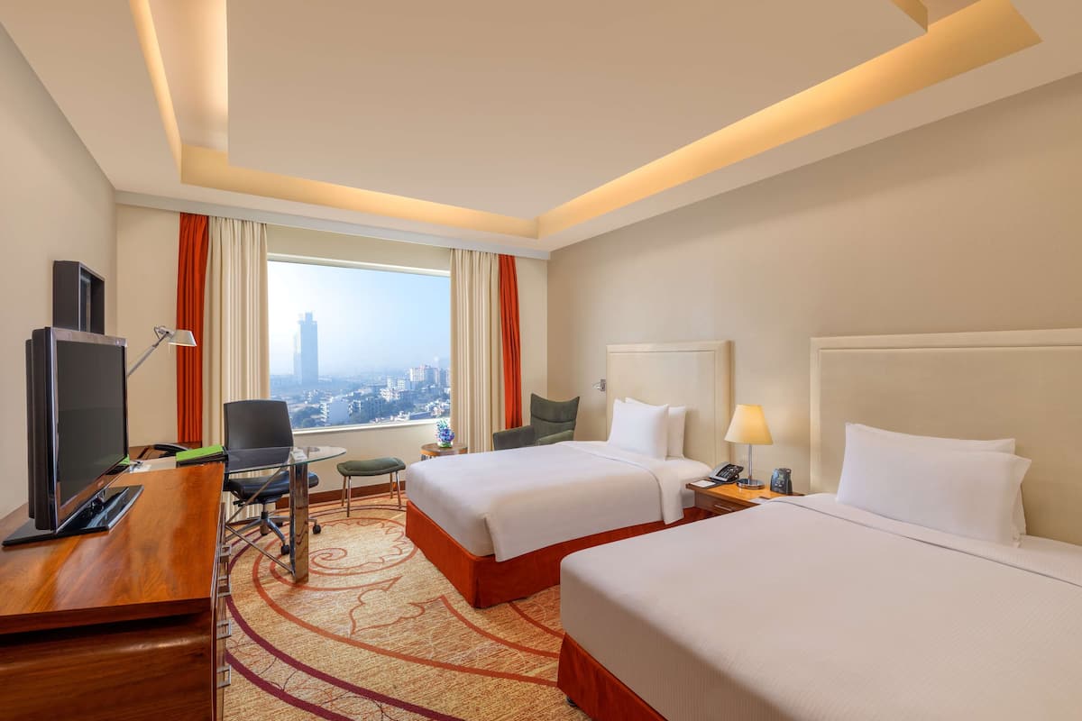 A hotel room with two beds, a desk with a TV, a chair, a small table, and a view of a cityscape.