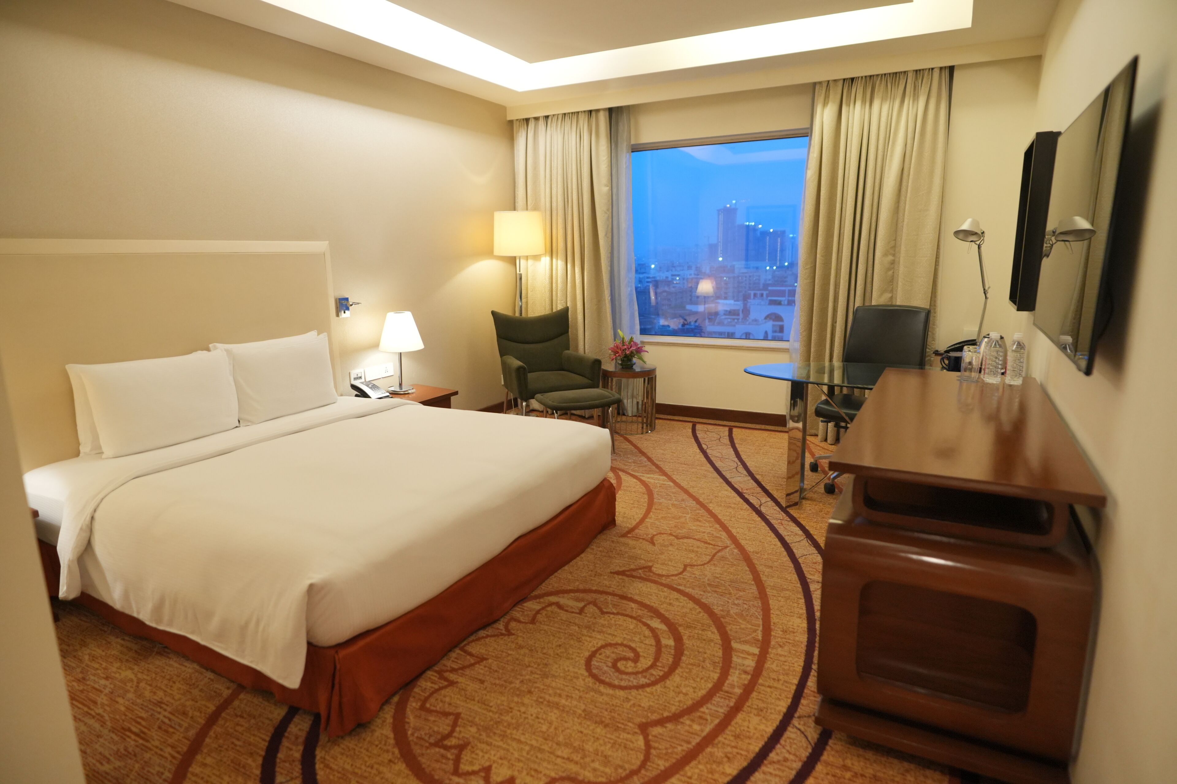 A hotel room with a large bed, a desk with a chair, a TV, and a view of the cityscape through the window.