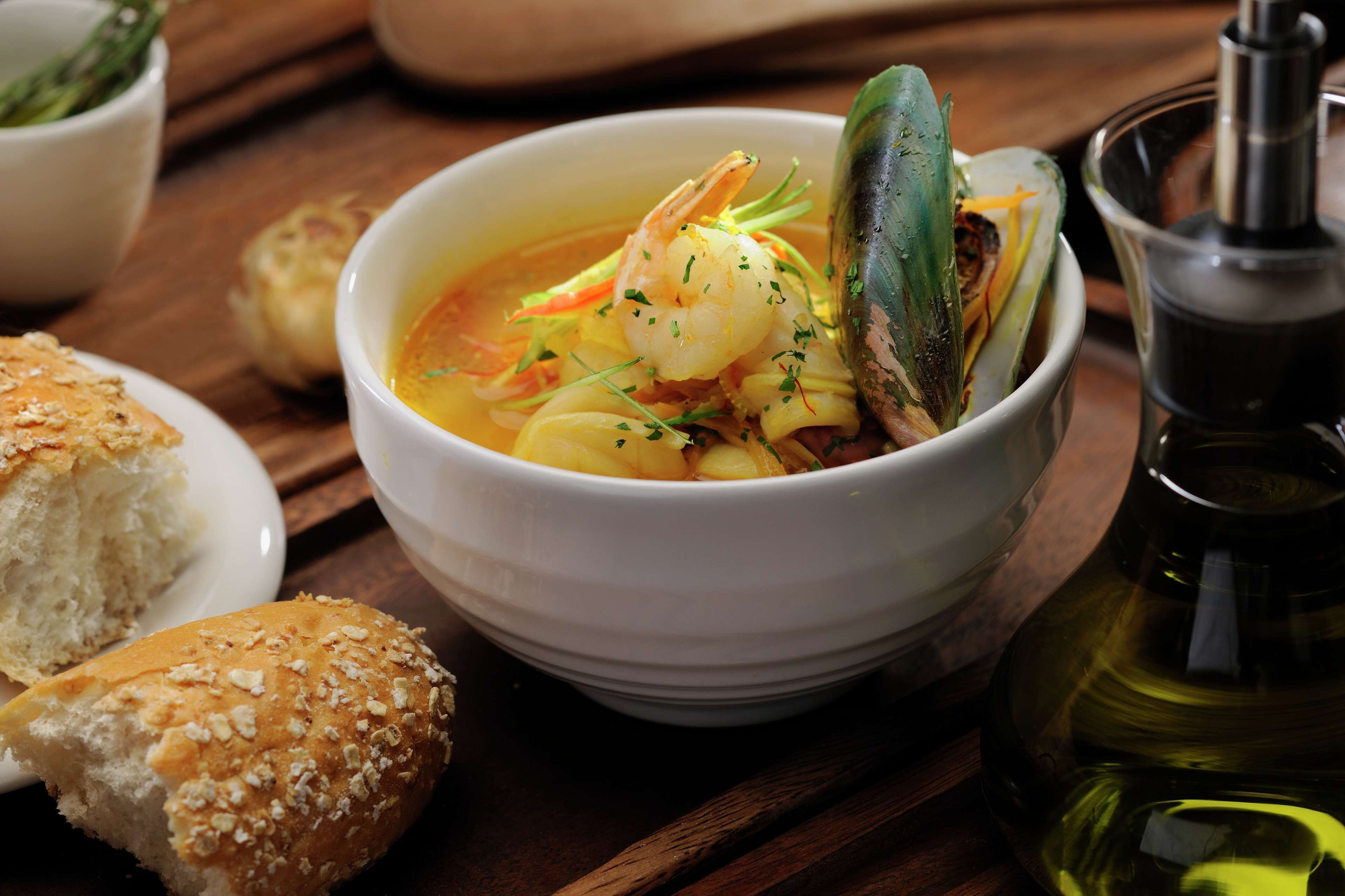 A bowl of seafood soup with bread and a bottle of oil on a wooden table.