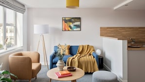A modern living room with a sofa, armchair, and coffee table, a painting on the wall, and a kitchen area with a wooden countertop.