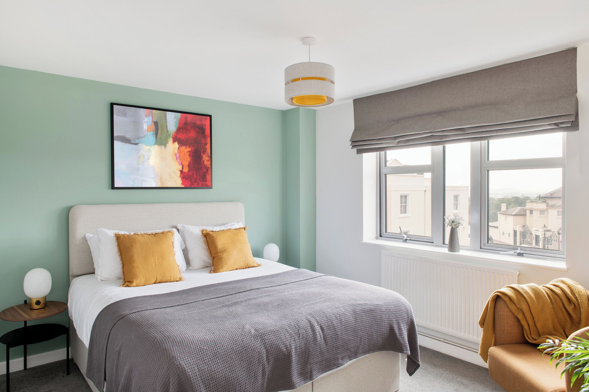 A bedroom with a bed, a bedside table, a lamp, a window with a view, and a painting on the wall.