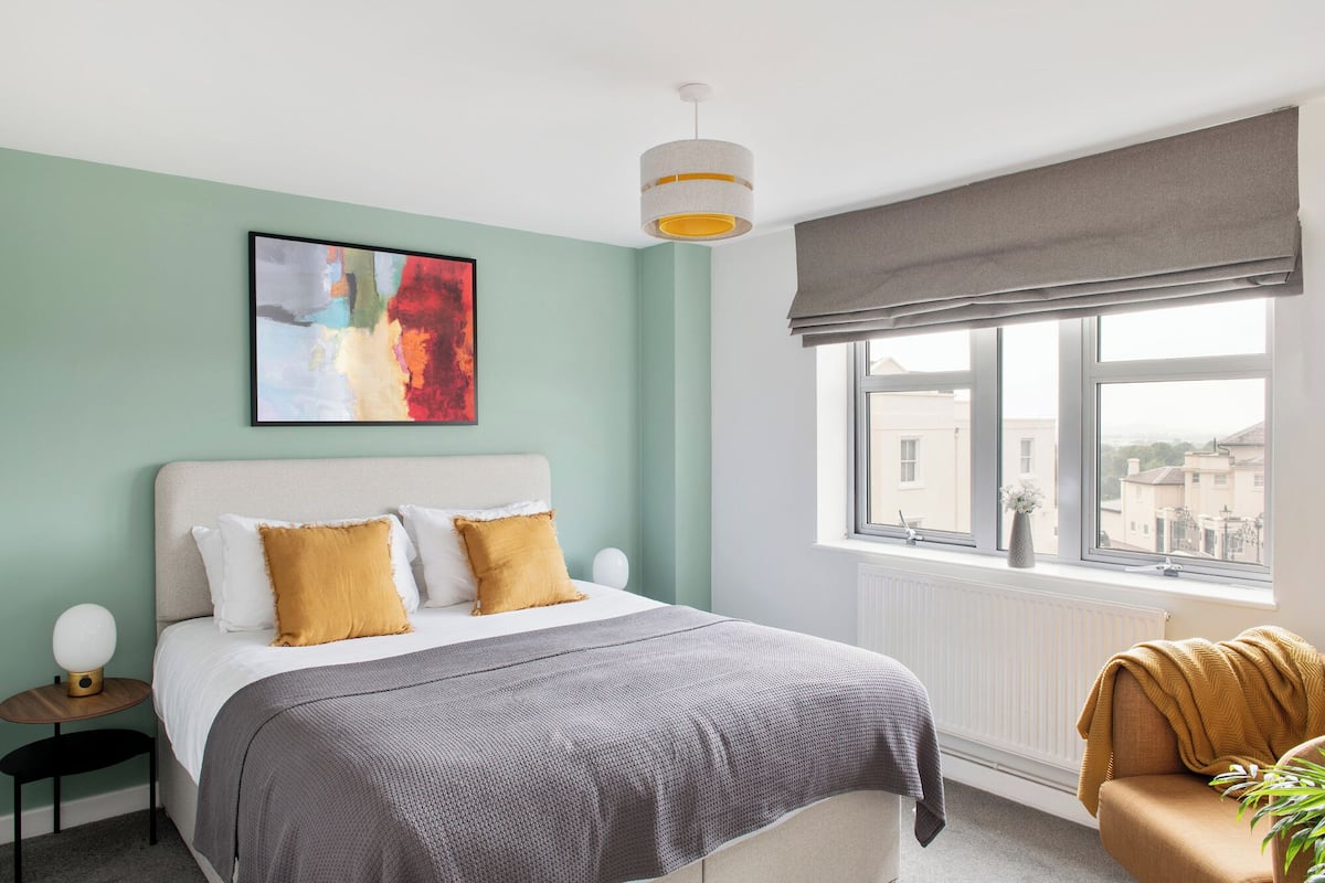 A bedroom with a bed, a bedside table, a lamp, a window with a view, and a painting on the wall.