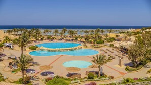 Ein Resort mit einem großen zentralen Pool, mehreren kleineren Pools in der Umgebung und einer Strandfläche mit Liegestühlen und Sonnenschirmen.