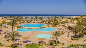 Un complejo turístico con una gran piscina central, piscinas más pequeñas alrededor y una zona de playa con tumbonas y sombrillas.