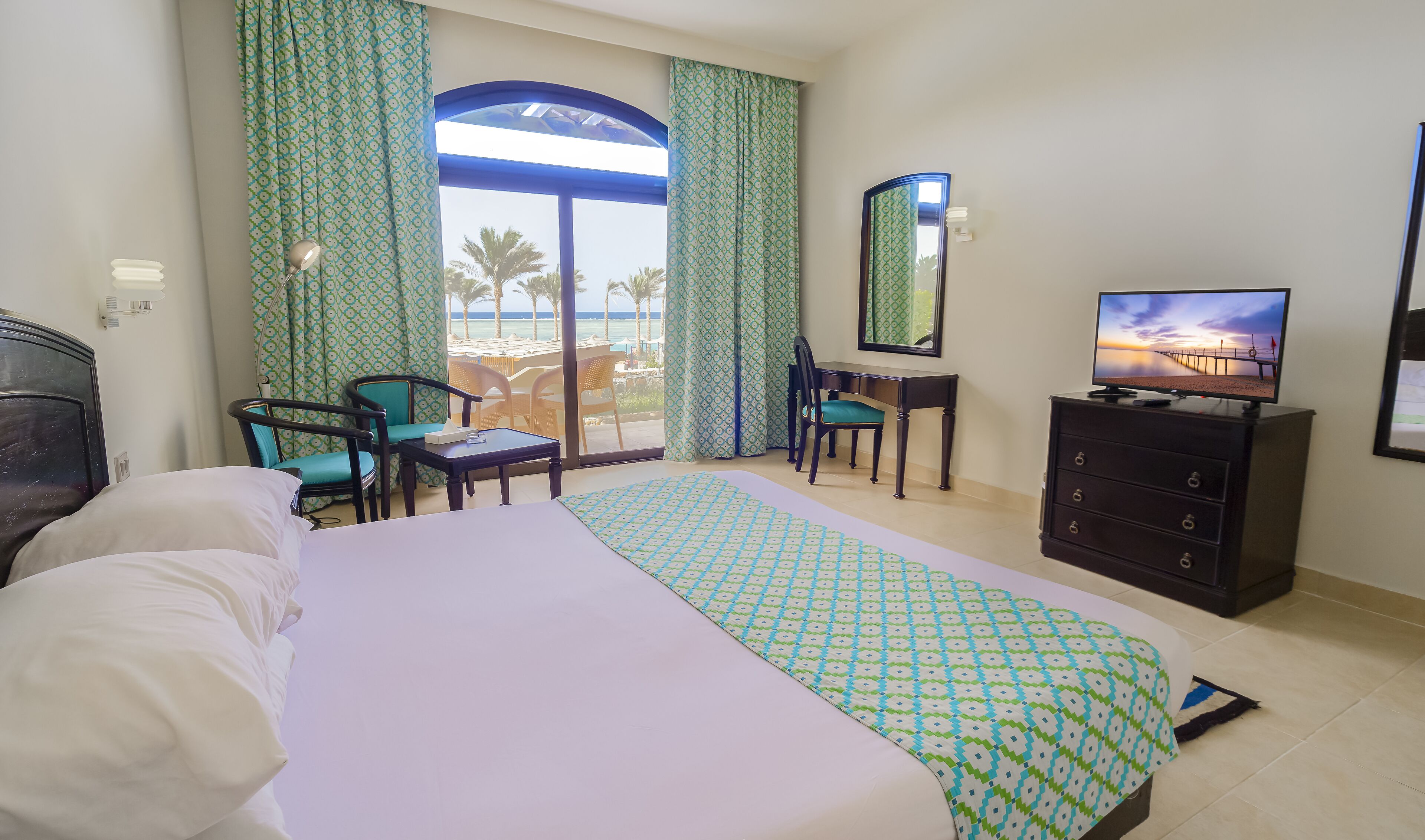 A hotel room with a bed, a desk, a chair, a television, and a view of the beach.