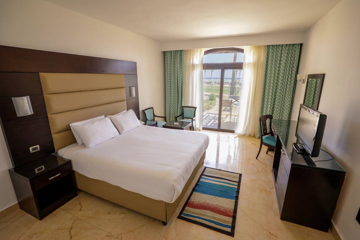 A hotel room with a large bed, a nightstand, a desk with a computer, a TV, and a balcony with a view.