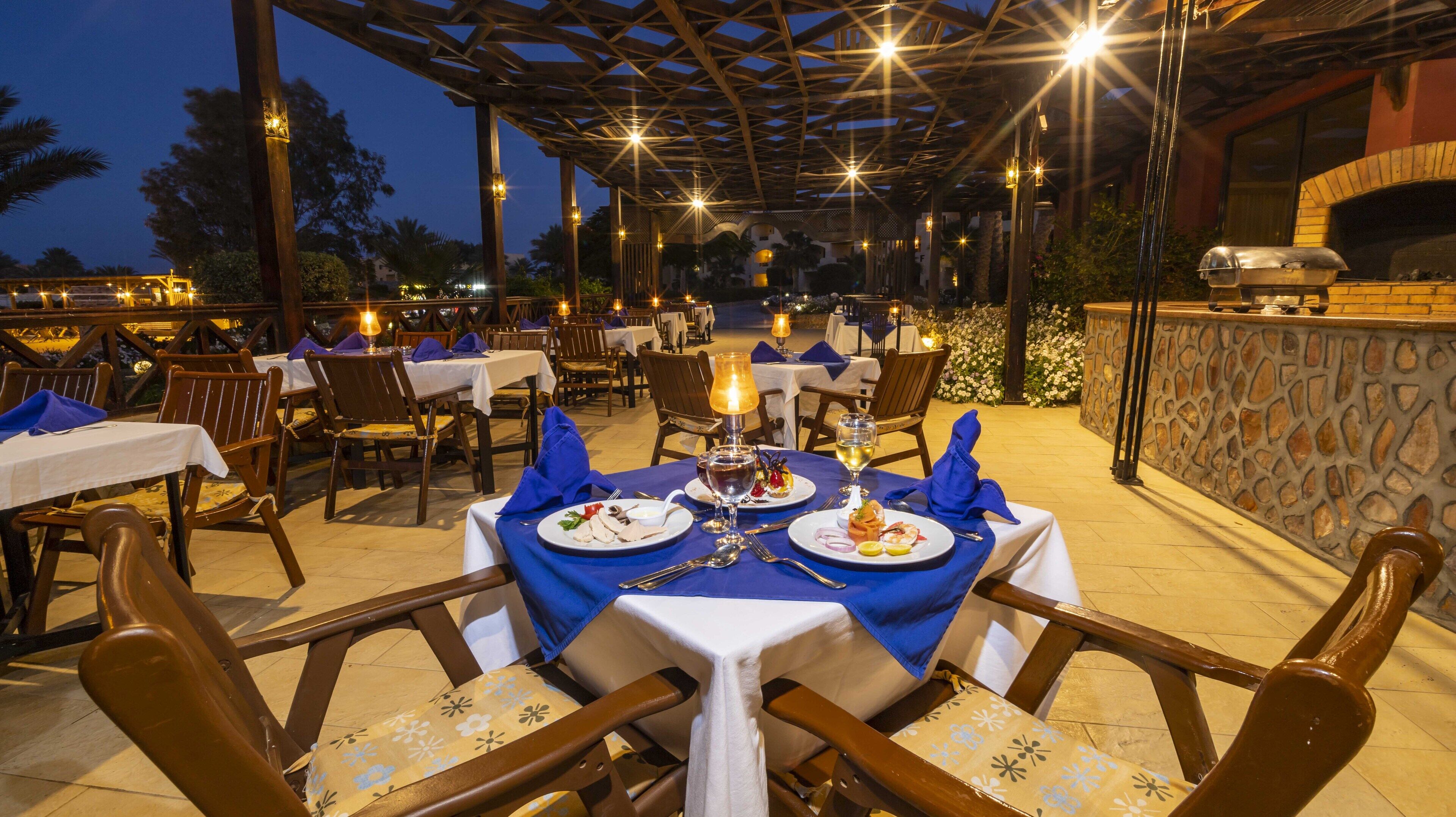 Un área de comedor al aire libre con mesas dispuestas para dos, bajo una pérgola de madera con luces colgantes.