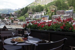 A table set with a bottle of wine, glasses, and a plate of food, overlooking a town with red flowers and buildings. - Schweizerhof Zermatt (Zermatt)
