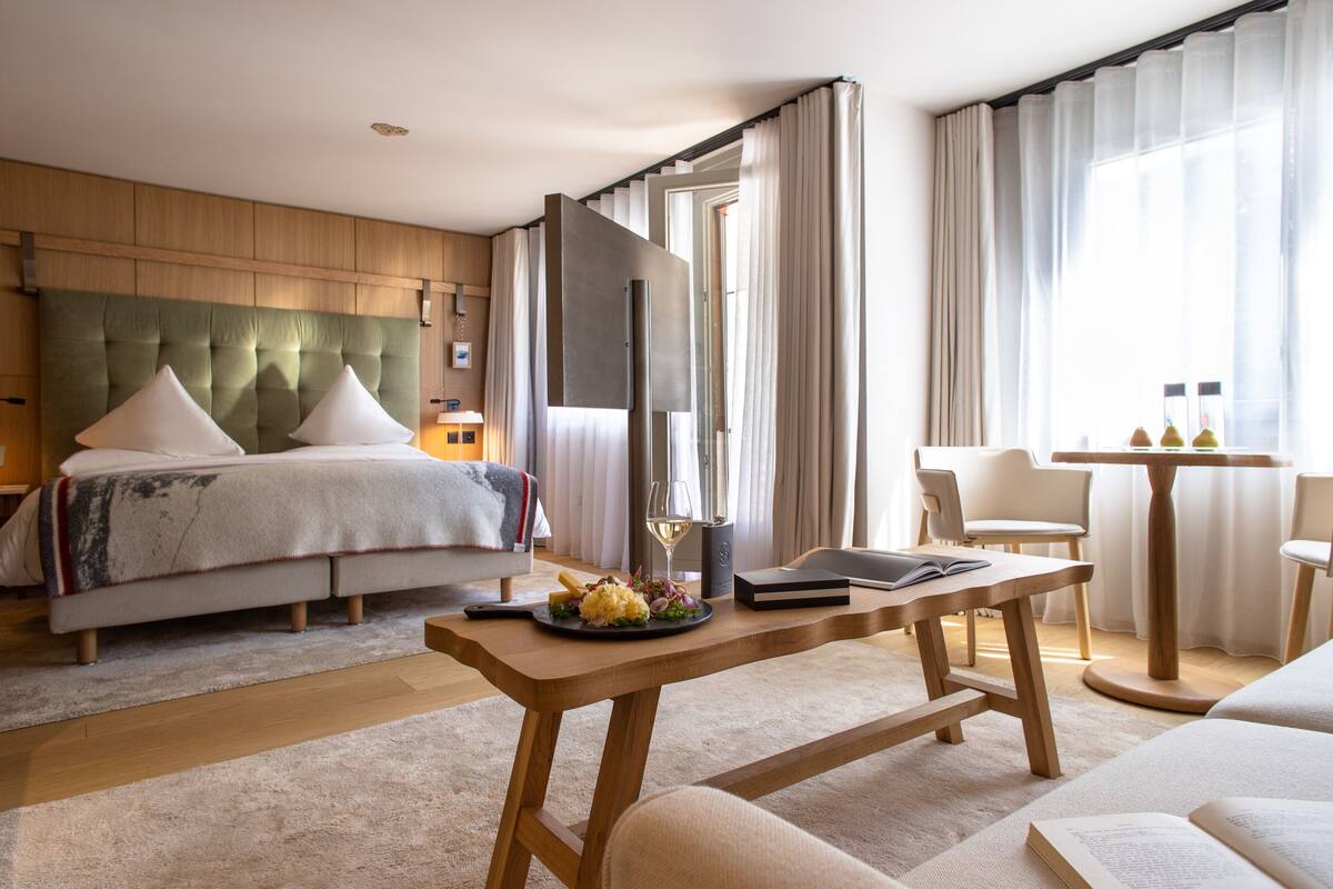 A modern hotel room with a large bed, a wooden headboard, a dining table with a plate of food, a book, and a TV.