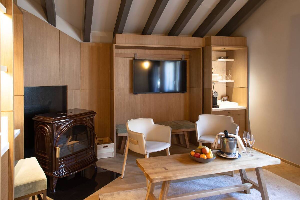 A modern living room with a wooden TV stand, two white chairs, a wooden table with fruit, and a built-in shelf with glassware.
