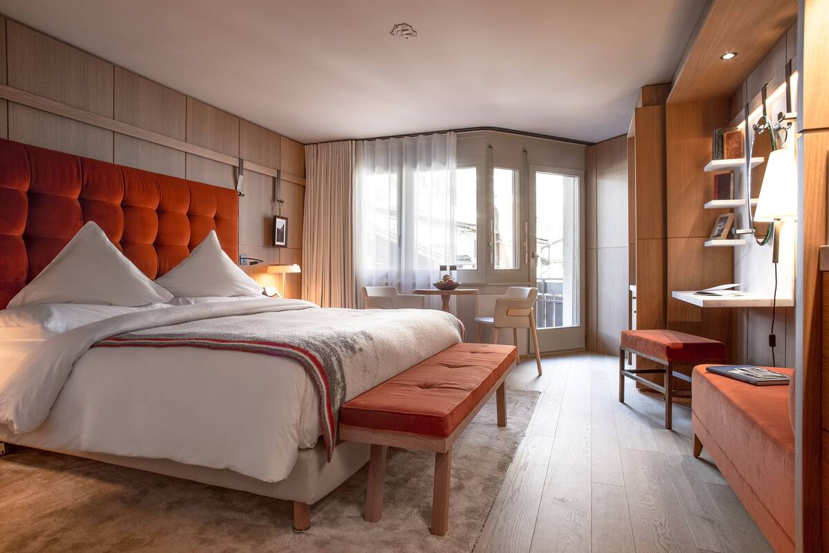 A modern bedroom with a tufted headboard, a large bed with white linens, a wooden bench, a desk, and a bookshelf.