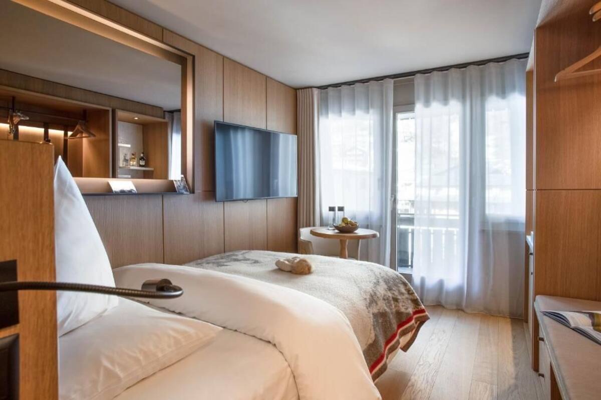 A hotel room with a large bed, a flat-screen TV mounted on the wall, a small round table, and a window with curtains.